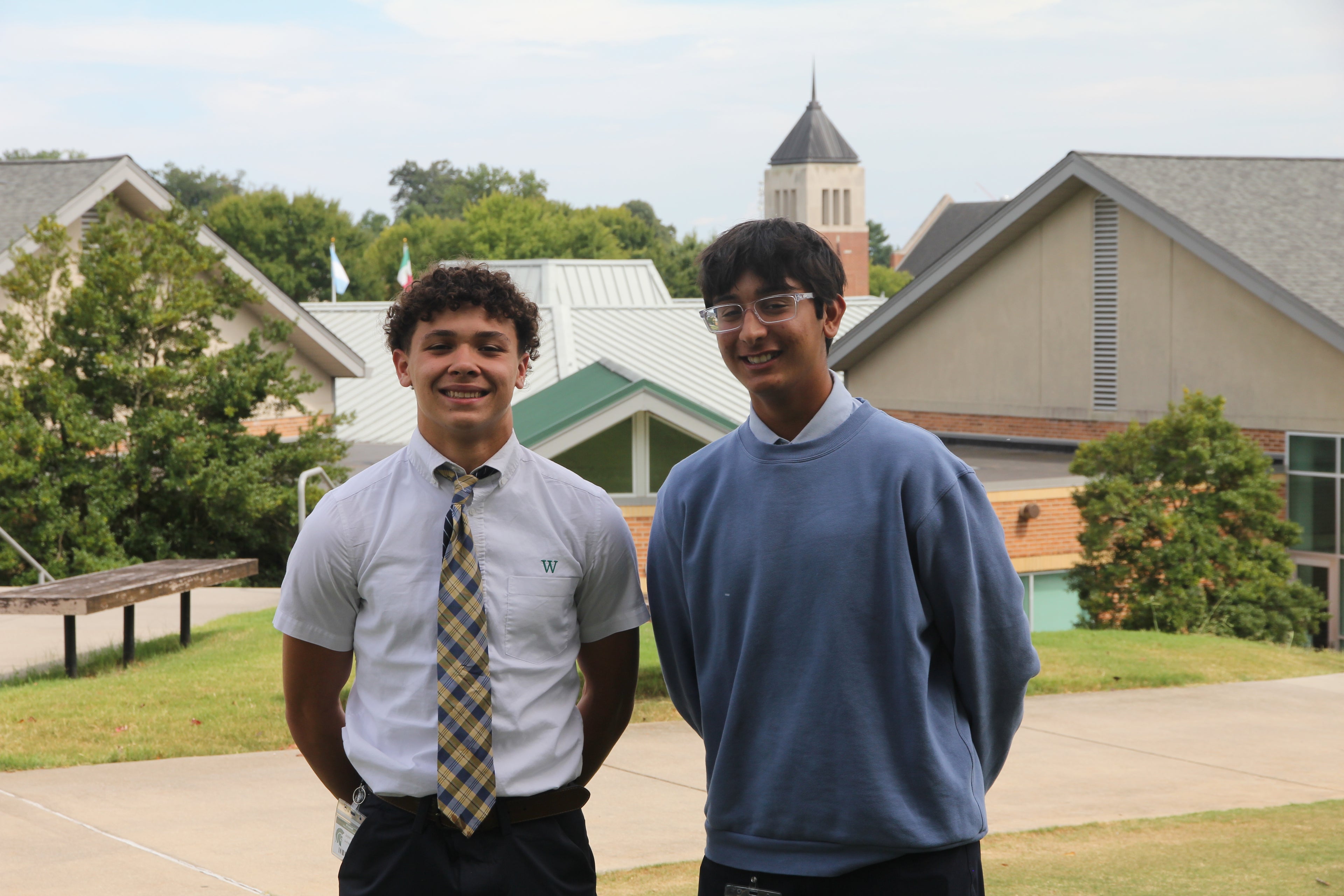 Two men standing next to each other, smiling, school building and trees in background. blue sky.